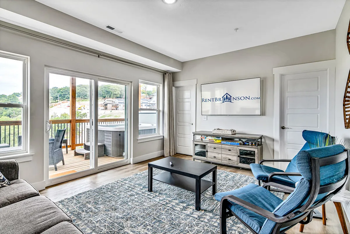 A modern, cozy lower-level living area featuring a comfortable sofa, blue accent chairs, a coffee table, large windows letting in natural light, and a wall-mounted TV—ideal for gatherings or enjoying movies.