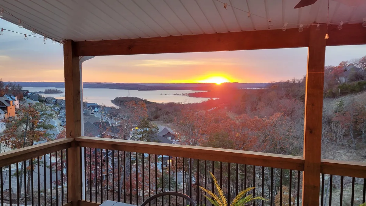 The hero image features a tranquil sunset casting golden hues over Table Rock Lake, framed by lush, tree-lined hills. The vantage point is from a spacious porch, ideal for relaxing, entertaining, and soaking in the serene lakeside beauty. Distant houses dot the landscape, adding a sense of peaceful seclusion and exclusivity.
