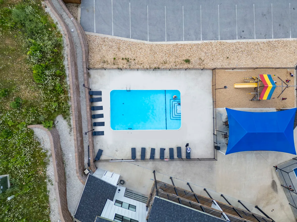 An inviting outdoor community pool area featuring lounge chairs, a blue shade structure, and a visible children’s play area. The mood is bright and cheerful, suggesting family fun and relaxation under the sun.