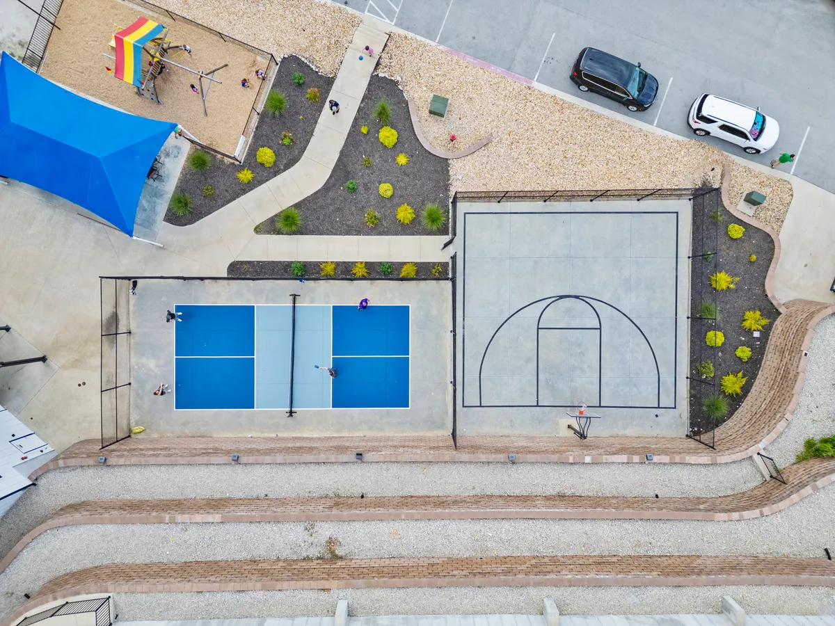 A dynamic exterior view of a well-maintained community multi-sport court, featuring basketball and tennis courts, a playground area, and landscaped gardens. People can be imagined playing or gathering, creating a sense of communal activity.