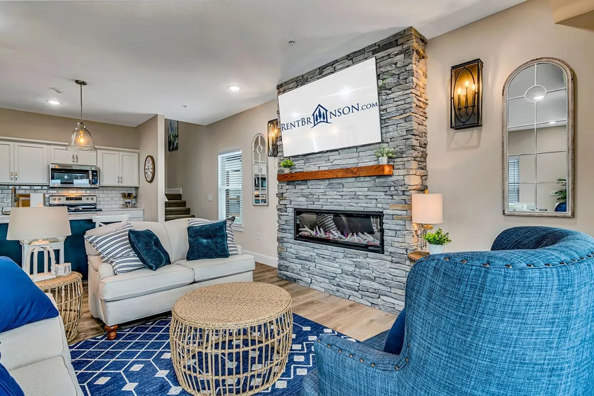 A cozy living room centers around a striking stone fireplace, flanked by blue accent chairs and a woven coffee table. In the background, a sleek, modern kitchen area is visible, reinforcing the open-concept design ideal for entertaining family and friends.