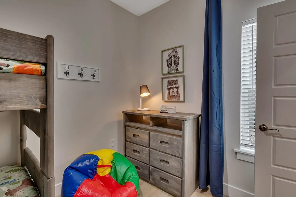 A cheerful, kid-friendly bunk room with a sturdy bunk bed, a colorful bean bag chair, a small dresser topped with a lamp, and whimsical framed animal artwork on the walls. The mood is playful and inviting—ideal for children.