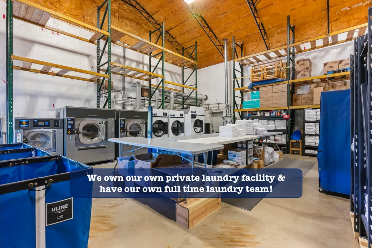A busy and organized commercial laundry facility featuring industrial washing machines, a laundry folding table, storage shelves, and laundry bins. The space is clean and efficient, representing a professional approach to cleanliness.