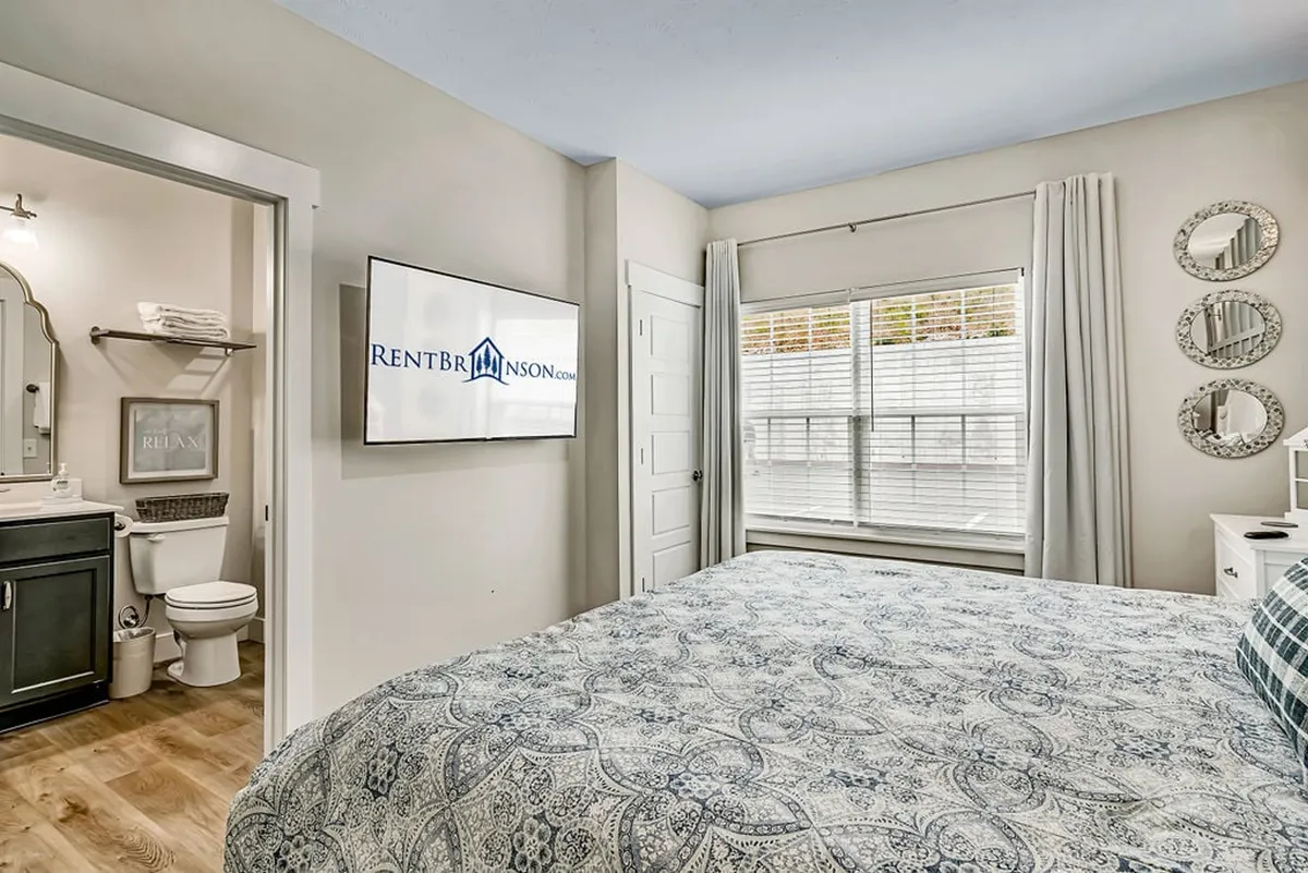 A spacious bedroom featuring a large bed with patterned bedding, a wall-mounted television, a window letting in soft natural light, and a doorway leading to a modern ensuite bathroom. The space feels calm, comfortable, and private.