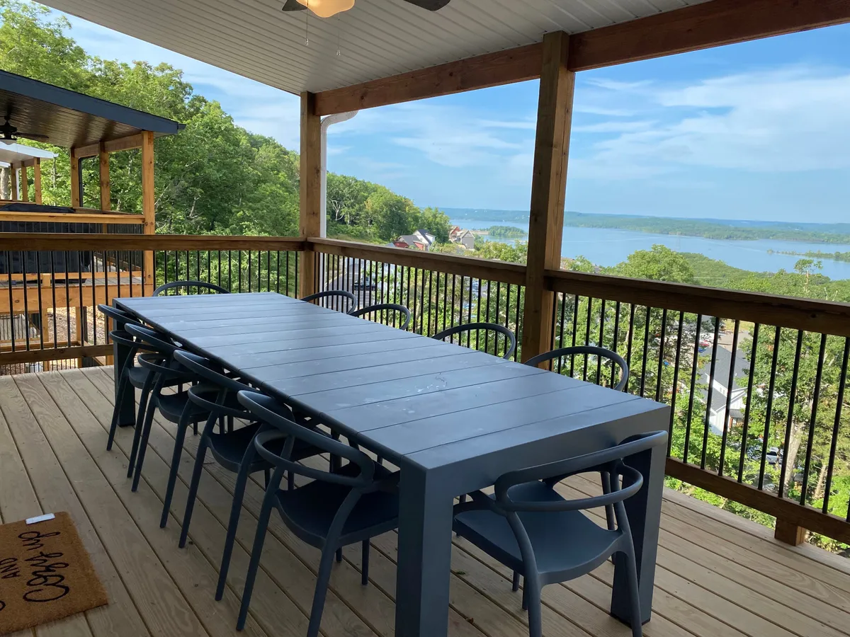 This image captures a large outdoor dining table on a scenic deck overlooking the lake, with a wooden railing framing the vista. The setting suggests ample space for relaxing, dining, and enjoying the outdoors together, with the tranquil lake providing a picturesque backdrop.