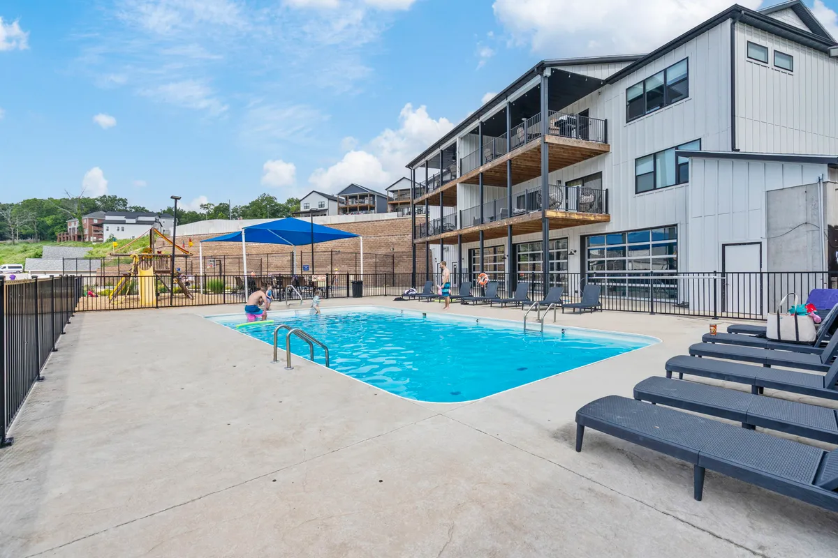 A cheerful exterior scene featuring a sparkling outdoor swimming pool surrounded by lounge chairs, the building facade in the background, and a children’s play area under a shade structure. The setting is lively, suggesting summer fun and social interaction.