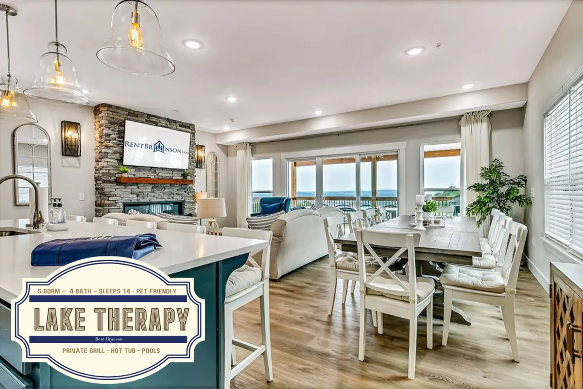 A spacious, open-plan living area combines a modern kitchen with stainless steel appliances, a generous island, an elegant dining table, large windows framing a scenic view, and a cozy stone fireplace. The layout is ideal for both cooking and gathering, with seamless visual flow between the kitchen, dining, and living room.