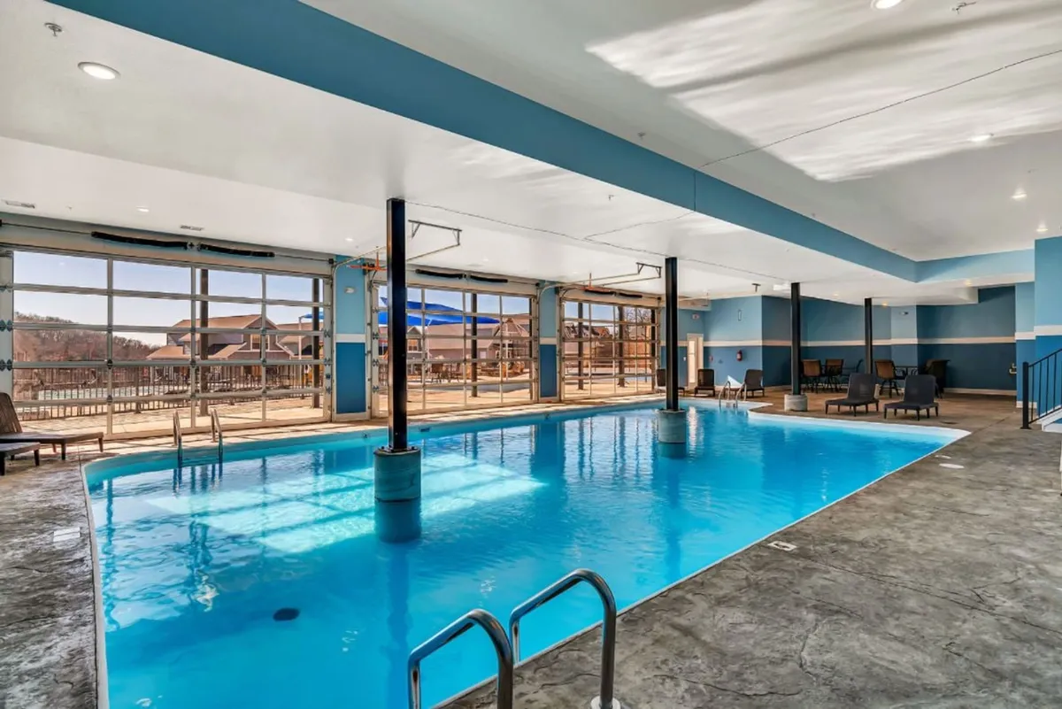 An elegant indoor pool area is bathed in natural light from floor-to-ceiling windows, with lounge chairs lining the poolside. The environment is tranquil and upscale, suggesting a resort-style amenity available to guests.