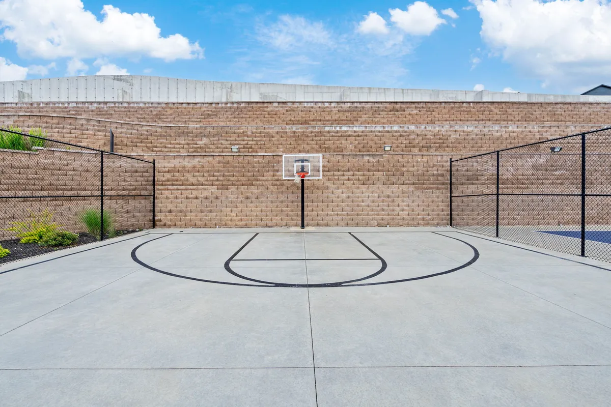 An active exterior scene featuring a smooth multi-sport court with a basketball hoop, surrounded by a chain-link fence. The space is inviting for sports, group games, and friendly competition, ideal for both adult and family play.